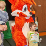 Young boy holds his Imagination Graduation diploma, smiles, and poses with Booker the Fox mascot