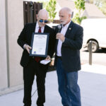 Man speaks into microphone. Karl holds framed paper.
