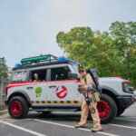 Teen dressed up as a ghostbuster poses in front of ghostbuster jeep